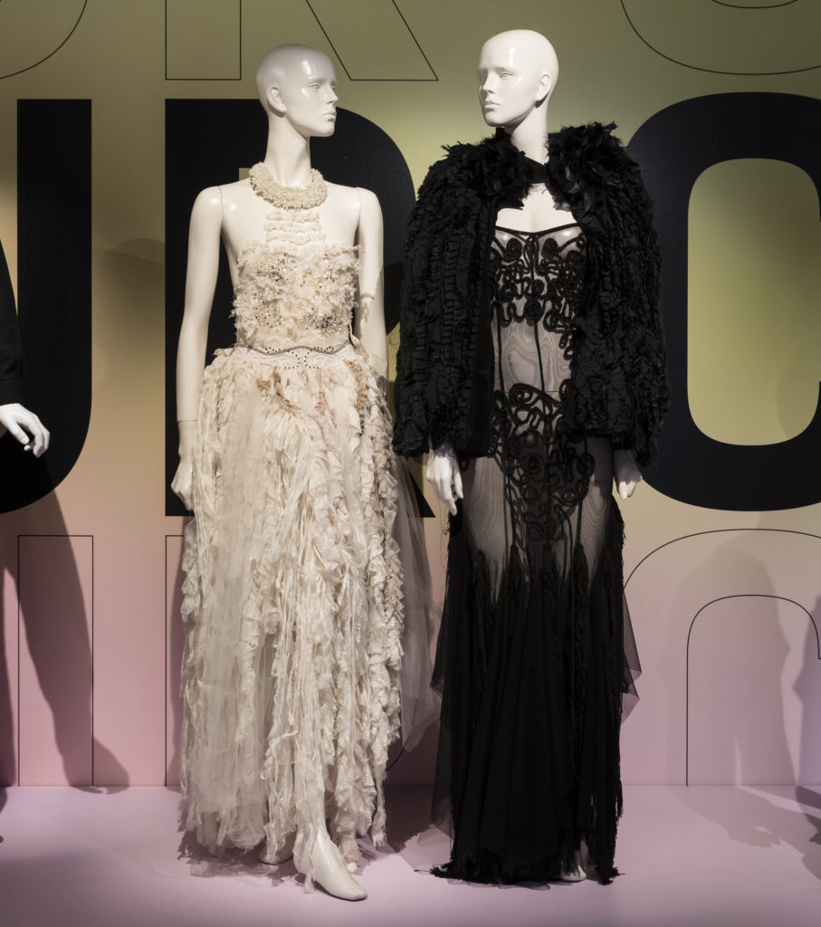 Two mannequins on display, one wearing white dress, the other wearing sheer black dress and cape.